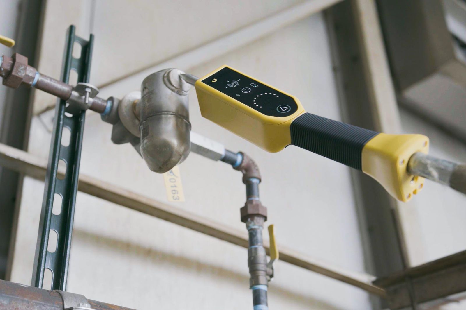 A handheld ultrasonic tester held against a steam trap on overhead pipework, with a numbered inspection tag on the trap.