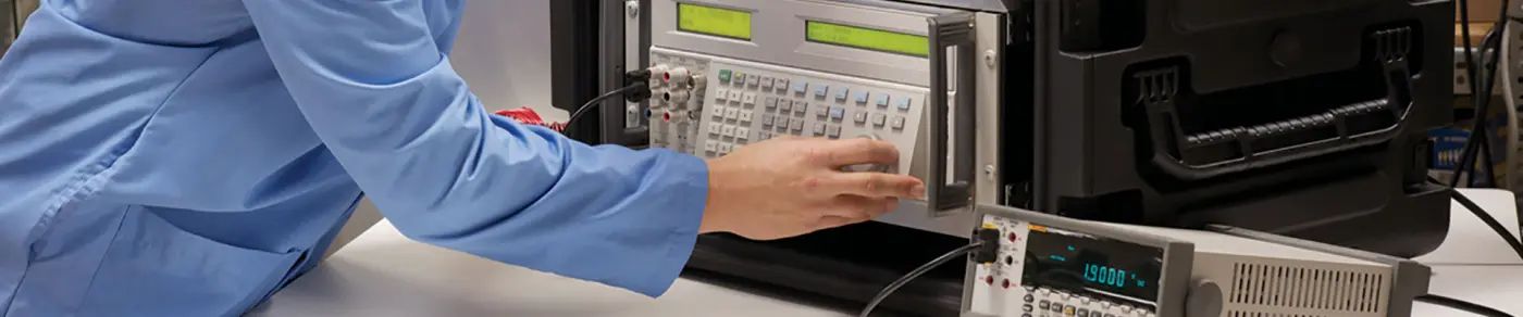 A technician in a lab coat operating a rack-mounted calibrator on a bench, beside a digital meter and a hard transit case.