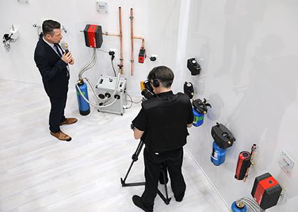Wall-mounted water filtration and treatment units with copper pipework in a showroom, being filmed.